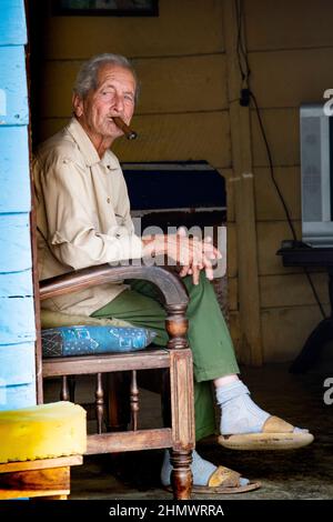 Uomo più anziano che fuma un sigaro cubano mentre si siede su una sedia sul portico anteriore della sua casa a Trinidad, Cuba un sito patrimonio dell'umanità dell'UNESCO. Foto Stock