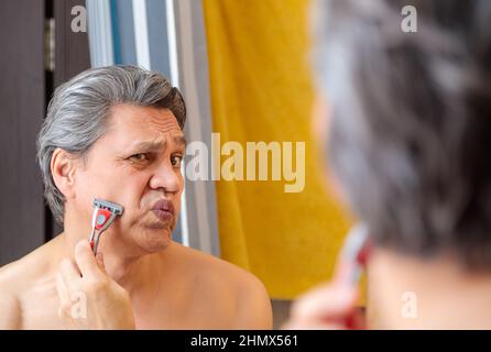 Un uomo adulto dai capelli grigi rasa con un rasoio davanti a uno specchio. Foto Stock