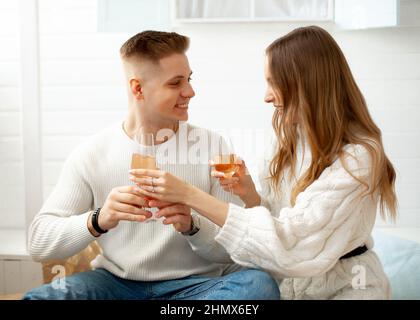 Donna dai capelli castani con capelli lunghi in abito a maglia bianco di maglia grande e carino giovane uomo sorridere l'un l'altro con bicchieri di vino nelle loro mani Foto Stock