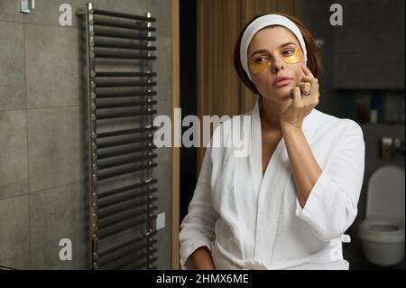 Bella donna incinta caucasica applicando cerotti di lisciamento di collagene dorato sotto gli occhi in bagno di casa. Rimuovere grinze, puffiness e cir scuro Foto Stock