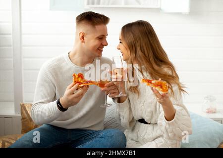 Il giovane uomo sorridente in maglione bianco e jeans ama il gusto della pizza e del bicchiere di vino con la sua ragazza, vestita in tunica bianca a maglia larga Foto Stock