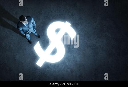 Vista dall'alto di un uomo d'affari in piedi davanti ad un simbolo grande del dollaro di soldi incandescente. Concetto di arricchirsi. Rendering fotorealistico 3D. Foto Stock