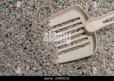 Primo piano della lettiera di gatto sbriciolata sotto forma di palline grigie, bianche e blu con una paletta di plastica bianca all'interno. Foto Stock