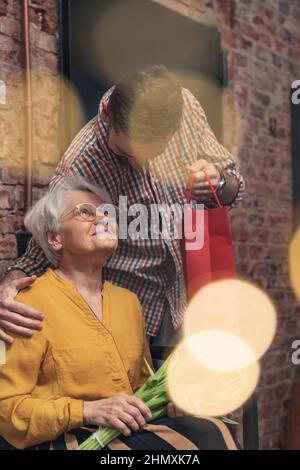 Stretta relazione tra la nonna in pensione dai capelli grigi che riceve un regalo dal suo nipote più vecchio il giorno del nonno. Foto di alta qualità Foto Stock