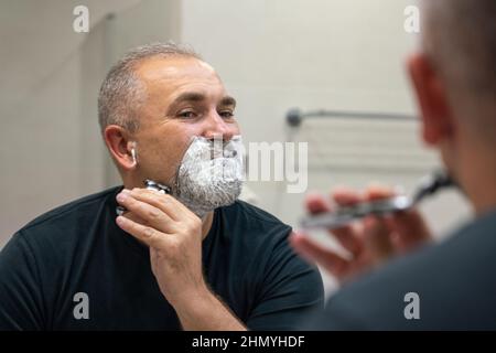 Bell'uomo barba dai capelli bianchi che rasa la barba guardando in uno specchio. Concetto di cambiare stile Foto Stock