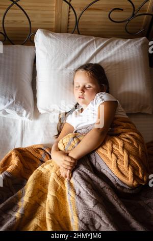 Giovane ragazza caucasica con pigtail che dorme in posizione attorcigliata su un grande letto matrimoniale con bocca leggermente aperta coperta da coperta gialla grigia Foto Stock