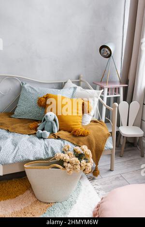 Un letto nella camera dei bambini con un giocattolo e cuscini. Accanto al letto si trova un cesto di vimini con fiori come elemento decorativo. Interni moderni Foto Stock