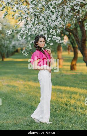 Una giovane donna carina in pantaloni bianchi e una blusa rosa brillante scorre attraverso un parco o giardino primaverile tra gli alberi fioriti di un albero di mele Foto Stock