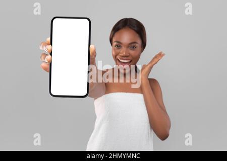 Mockup mobile. Happy Excited Black Lady avvolta in asciugamano che dimostra smartphone vuoto Foto Stock