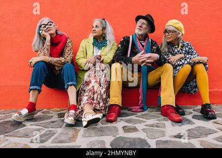 Persone anziane che guardano via con attenzione mentre si siedono insieme contro un muro rosso. Gli anziani pensivi riflettono sui ricordi del passato. Gruppo di matu Foto Stock