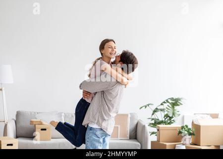 Soddisfatto europeo giovane marito solleva e abbraccia sua moglie in camera interni divertirsi insieme Foto Stock