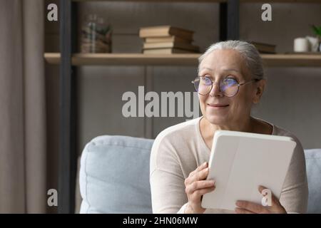 Sorridente utente di tablet anziano seduto sul divano con buoni pensieri Foto Stock