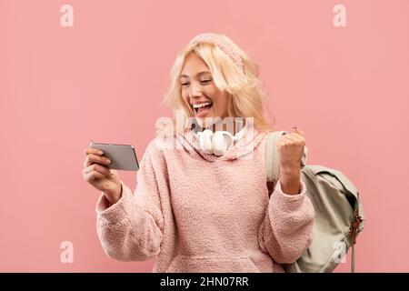 Una studentessa molto gioiosa che tiene smartphone e scuote il pugno in gioia, posando con zaino su sfondo rosa Foto Stock