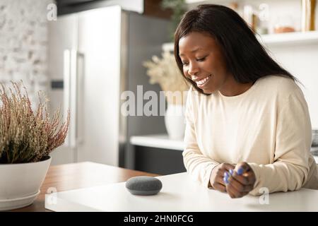 Sorridente donna afroamericana sta usando il dispositivo di assistente domestico intelligente con la funzione di riconoscimento vocale, signora multirazziale che parla con l'altoparlante intelligente portatile. Concetto di casa intelligente Foto Stock