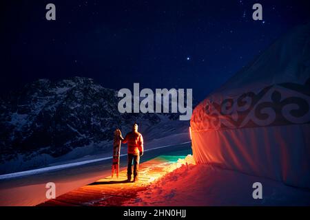 Uomo che tiene split snowboard vicino Yurt nomadic casa complesso presso la stazione sciistica di Shymbulak in Almaty, Kazakistan. Astrofotografia notturna invernale con stelle A. Foto Stock