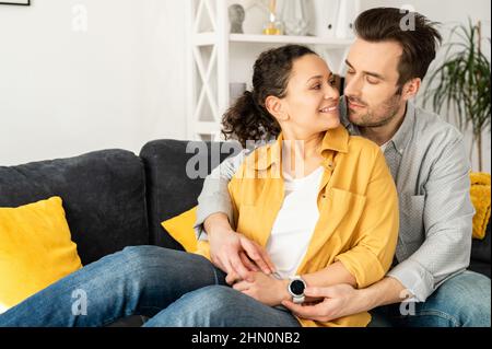 Coppia dolce e graziosa di mezza età innamorata seduta in abbraccio sul comodo divano a casa, che si unisce teneramente l'uno all'altro. Concetto di sostegno e cura nel matrimonio Foto Stock