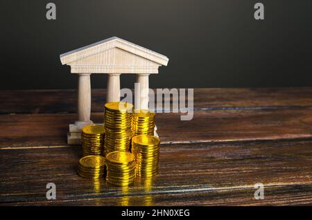 Edificio bancario e pile di monete. Debito statale. Budget. Riscossione delle imposte. Investimenti in istruzione, musei e biblioteche, istituzioni governative. Tappo banca Foto Stock