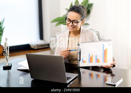 Dipendente multirazziale femminile durante una riunione virtuale, che mostra i grafici nella webcam del laptop, donna d'affari coinvolto conferenza, tutor o business coach leader webinar Foto Stock