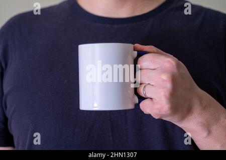 Uomo che tiene una tazza bianca di bevanda calda all'aperto. Mano dell'uomo che tiene una tazza bianca contro lo sfondo nero. Primo piano, immagine mockup, spazio di copia per il testo Foto Stock