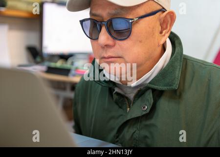 Un uomo che usa un portatile online indossando occhiali da sole e un cappello. Concetto di criminalità informatica, sicurezza, truffe finanziarie. Foto Stock