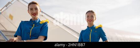 Due bellissime camere d'aria in uniforme blu che sorridono alla telecamera, in piedi su un'aerostazione e accogliendo i passeggeri Foto Stock