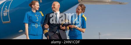 Eccitato pilota con due attraenti stewardesses in piedi insieme davanti ad un aeroplano e sorridente dopo l'atterraggio Foto Stock