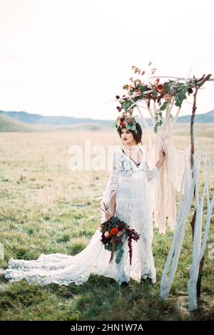 La sposa in una corona con un bouquet si erge su un tappeto bianco vicino a  un arco nuziale