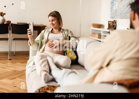 Donna incinta che prende selfie sul cellulare mentre si siede al divano in casa Foto Stock