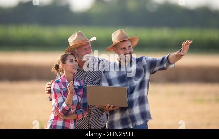 Agricoltore maturo con laptop che spiega qualcosa a una giovane agricoltore e a un anziano sul campo durante il raccolto in estate Foto Stock