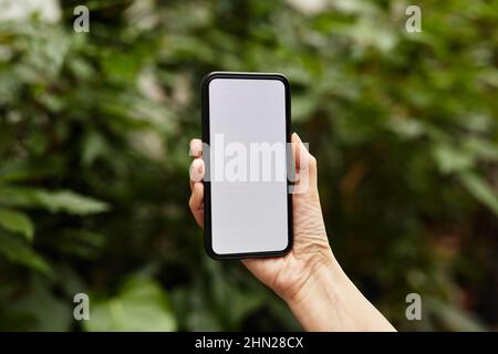 Primo piano di mano femminile che tiene smartphone contro la vegetazione verde durante il lavoro in serra moderna grande o orangerie Foto Stock