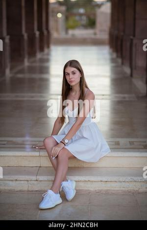 Una giovane bella donna in abito bianco siede su una scala di marmo Foto Stock