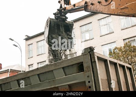 Lavori stradali sulla strada della città. La benna dell'escavatore raccoglie la vecchia pavimentazione e la carica in un dumper. Foto Stock