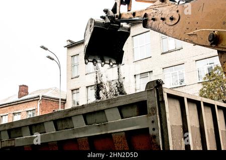 Lavori stradali sulla strada della città. La benna dell'escavatore raccoglie la vecchia pavimentazione e la carica in un dumper. Foto Stock