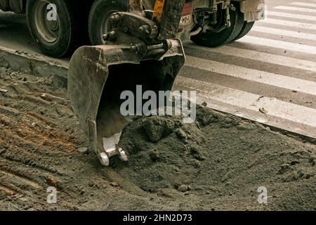 Lavori stradali sulla strada della città. La benna dell'escavatore raccoglie la vecchia pavimentazione e la carica in un dumper. Foto Stock