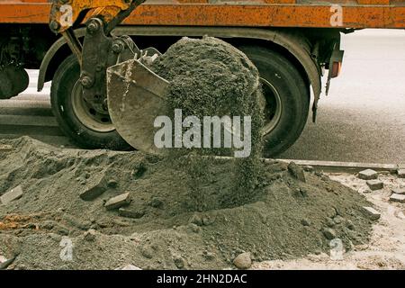 Lavori stradali sulla strada della città. La benna dell'escavatore raccoglie la vecchia pavimentazione e la carica in un dumper. Foto Stock