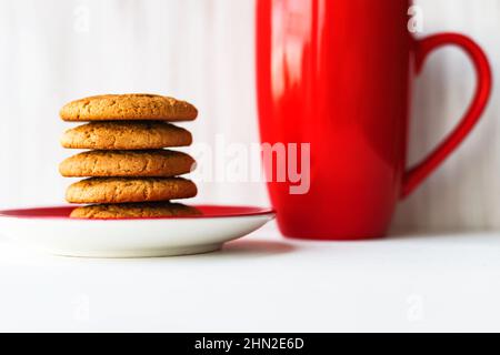 Accatastare i biscotti di farina d'avena sul piatto rosso con la tazza di caffè sul tavolo di legno bianco sullo sfondo della parete Foto Stock