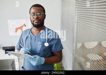 Giovane sicuro veterinario afroamericano che fa note in documento medico e guardando la macchina fotografica in ospedale animale Foto Stock