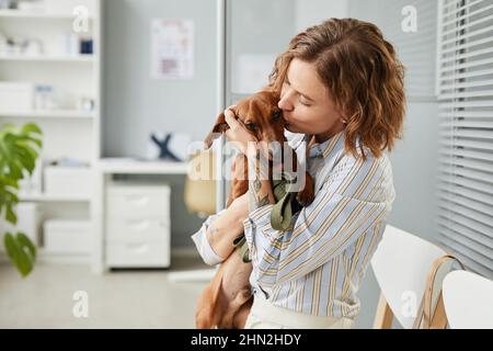 Giovane donna contemporanea che dà il bacio al dachshund marrone mentre tiene il suo animale domestico carino sulle mani nella sala dell'ospedale veterinario Foto Stock