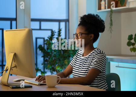 Giovane donna africana felice telelavoratore indossare occhiali di battitura a macchina, godendo di lavoro freelance Foto Stock