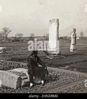 Foto d'epoca dell'uomo cinese in abito tradizionale e con coda seduta nell'orto in un cimitero della dinastia Ming. Cina settentrionale. 1906 Foto Stock