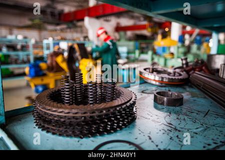 Foto ravvicinata delle parti metalliche della trasmissione con la molla su una tavola metallica incrinata. Stabilimento di riparazione autocarri. Bassa profondità di campo, parzialmente sfocata. Lavoratore su Foto Stock