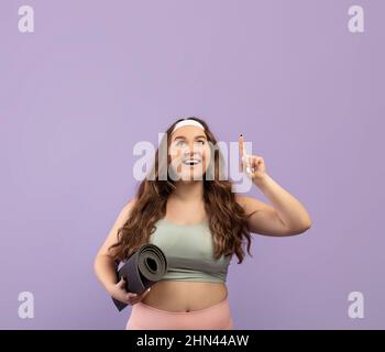 Felice divertente giovane europea Plus taglia donna in sportswear tenere il tappeto, mostrare il dito in su sullo spazio copia Foto Stock