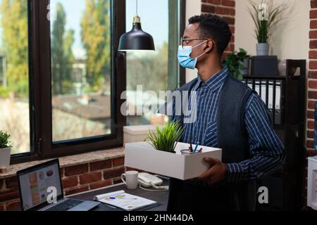 Dipendente afroamericano con maschera che osserva deluso fuori il vassoio di tenuta della finestra con gli effetti personali dopo essere stato sparato. Il lavoratore di startup perde lavoro dopo il ridimensionamento dell'azienda. Foto Stock