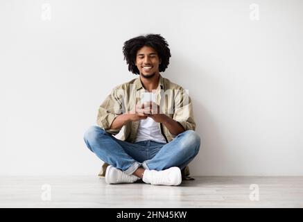 Comunicazione online. Messaggi Happy Black Man sullo smartphone mentre ci si rilassa sul pavimento Foto Stock