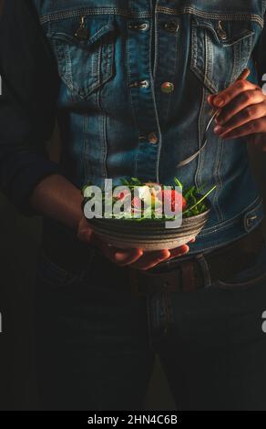 Pranzo vegano sano. Donna in denim blu tiene una ciotola in ceramica con avocado, rucola, pompelmo e insalata di anacardi nelle sue mani. Comfort, pulito, vegetaria Foto Stock