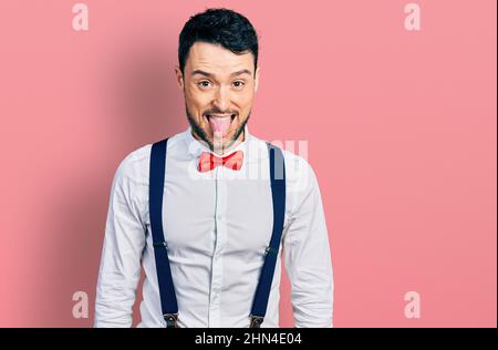 L'uomo ispanico con barba che indossa l'aspetto hipster con la cravatta di prua e sospenditori che attaccano la lingua fuori felice con espressione divertente. Concetto di emozione. Foto Stock