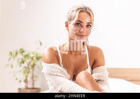 Una ragazza che guarda bene ha sul suo prodotto di pulizia della pelle del viso, seduta sul letto nella camera da letto. Foto Stock