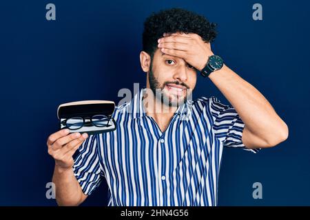 Giovane uomo arabo con barba che tiene i vetri in caso di occhiali stressati e frustrati con mano sulla testa, sorpreso e faccia arrabbiata Foto Stock