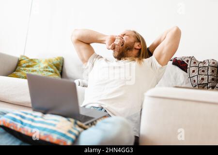 Biondo biondo biondo biondo biondo che sbava e usa il computer portatile mentre si siede sul pavimento di casa Foto Stock