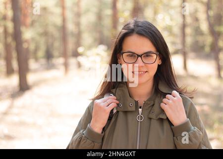 Primo piano ritratto di una bella ragazza sorridente con capelli lunghi e scuri in occhiali in piedi con uno zaino contro una foresta sfocata Foto Stock
